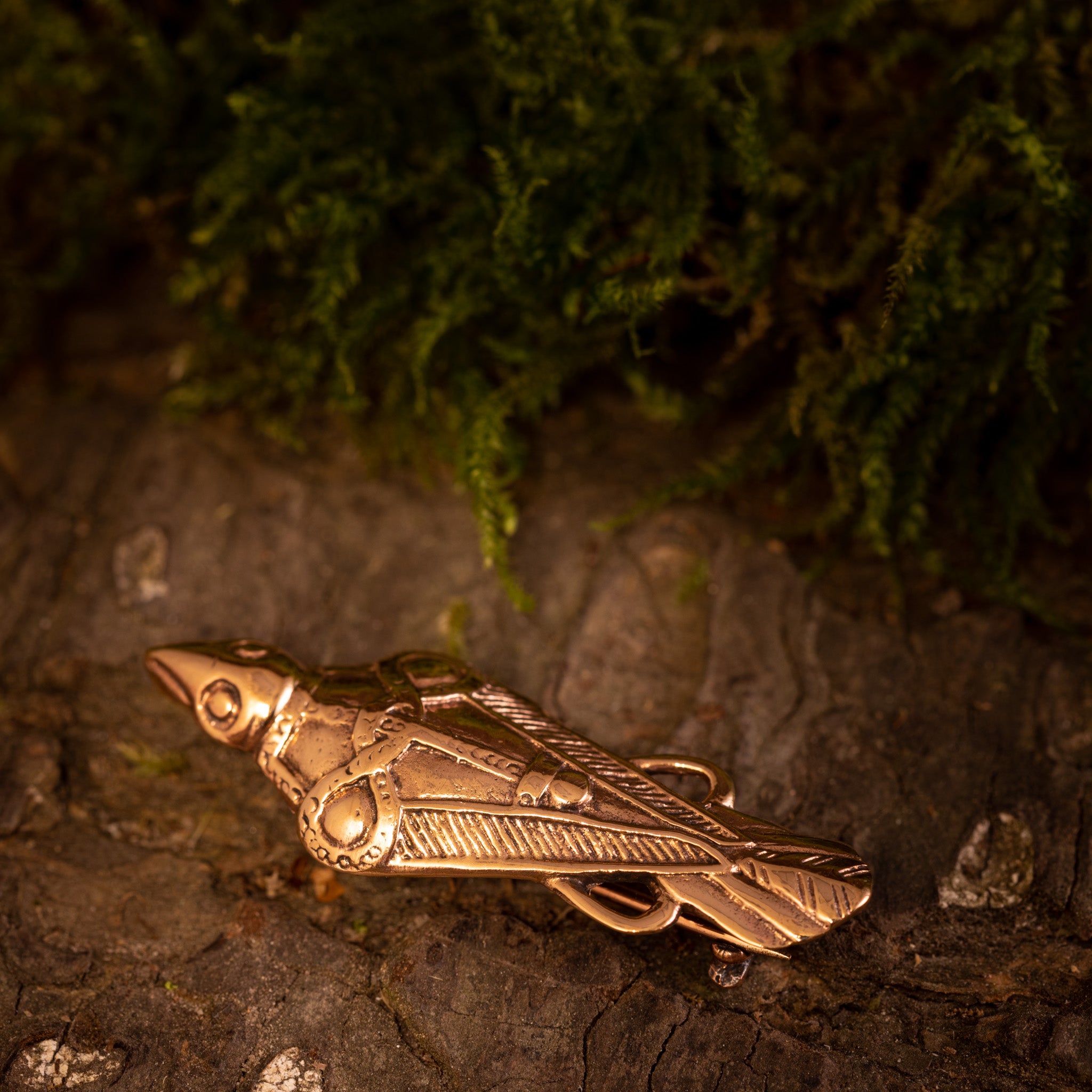 Broche Hugin / Munin Bronze, indviklet detaljeret med fugleformede graveringer, ligger på en rustik træoverflade med grønt mos i baggrunden. Denne antikke broche, der minder om Odins mytiske ravne, har en synlig låsemekanisme på ryggen.