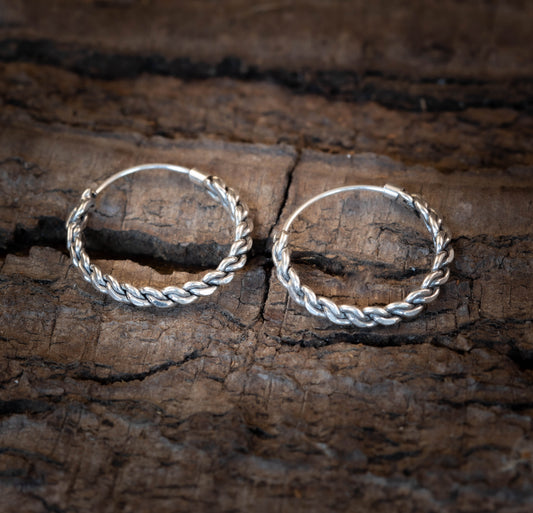 Ein Paar geflochtene 925s Creolen liegt auf einer strukturierten Holzoberfläche. Aus 925s Sterling Silver gefertigt, kontrastiert das kunstvolle Flechtdesign schön mit dem rustikalen Hintergrund.