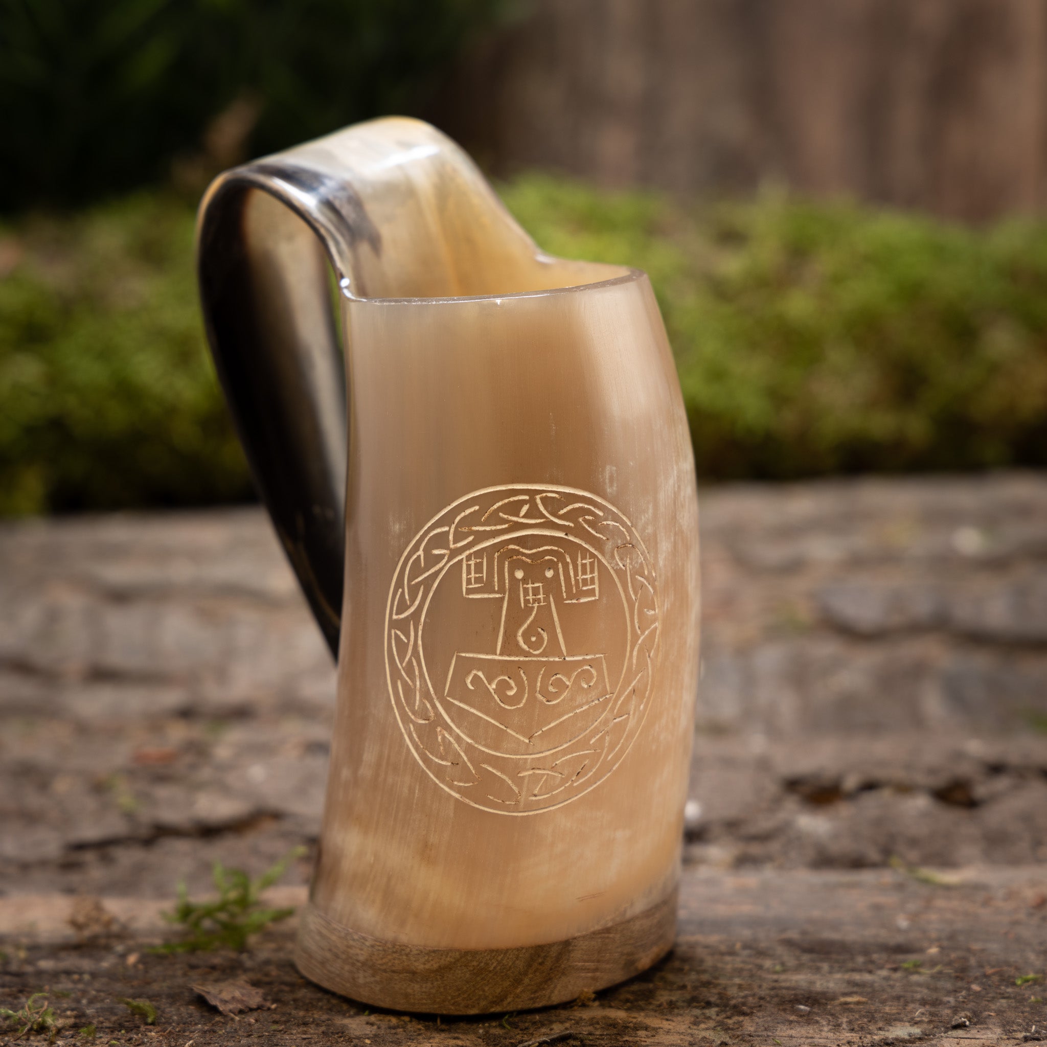 En Mini Vikingekrus Horn Thors Hammer (100 ml) sidder på en tekstureret stenoverflade, dens udsmykkede hornformede design minder om vikinge-drikkekar. Med et indgraveret cirkulært mønster med indviklede detaljer, muligvis forestillende et middelalder- eller fantasiemblem, kunne det nemt fremvises i Roskilde showroom. Baggrunden er sløret grønt og murværk.