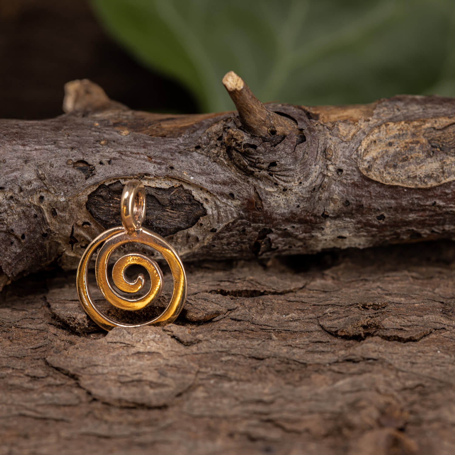 Et Spiral Vedhæng Bronze vedhæng med et indviklet gyldent design hviler på et tekstureret stykke bark. Den fremkalder ældgammel kunst og skiller sig ud mod det slørede grønne løv i baggrunden og understreger de naturlige og rustikke omgivelser, der minder om udstillingerne på Finlands Nationalmuseum.