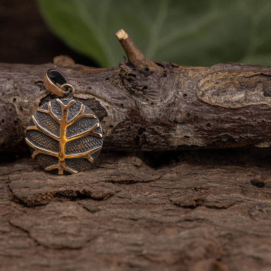 Yggdrasil Livets Træ Define Vedhæng Bronze, et rundt vedhæng med et detaljeret trædesign, ligger på en tekstureret træoverflade. Bronzemetallet står i kontrast mod den naturlige baggrund og fremhæver indviklede detaljer, der minder om nordisk mytologis Yggdrasil. Blødt slørede grønne blade kan ses i baggrunden.