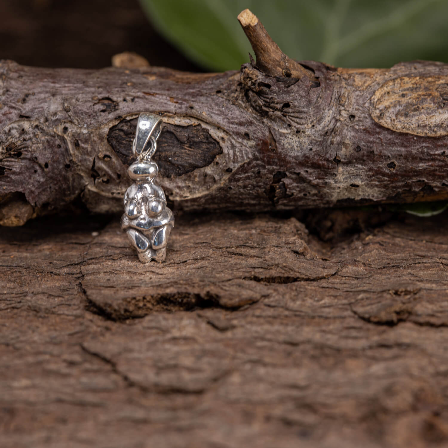 En lille Venus fra Willendorf Vedhæng 925s Sterling Sølv Lille, formet som en stiliseret sammenkrøbet figur, ligger på et stykke groft, struktureret træ. Baggrunden inkluderer grønne, ude af fokus blade, der fremhæver skærmens naturlige indstilling. Dette elegante tilbehør er lavet af 925s Sterling Sølv og udstråler tidløs charme.