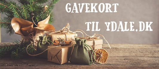 En rustik julescene med indpakkede gaver, grønne poser og tørrede appelsinskiver på en træoverflade. En lille stedsegrøn gren er i baggrunden. Teksten "Gavekort til ydale.dk - 300kr" vises tydeligt med hvide bogstaver.