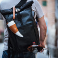 Bild in Galerie-Betrachter laden, En person iført en grå t-shirt bærer en stor sort roll-top rygsæk med en hvid og solbrun ged Kaffe Drikkehorn Brun påsat, mens han skubber en cykel med en brun Brooks-sadel i læder. Scenen fremkalder stemningen fra en moderne viking på en bygade.
