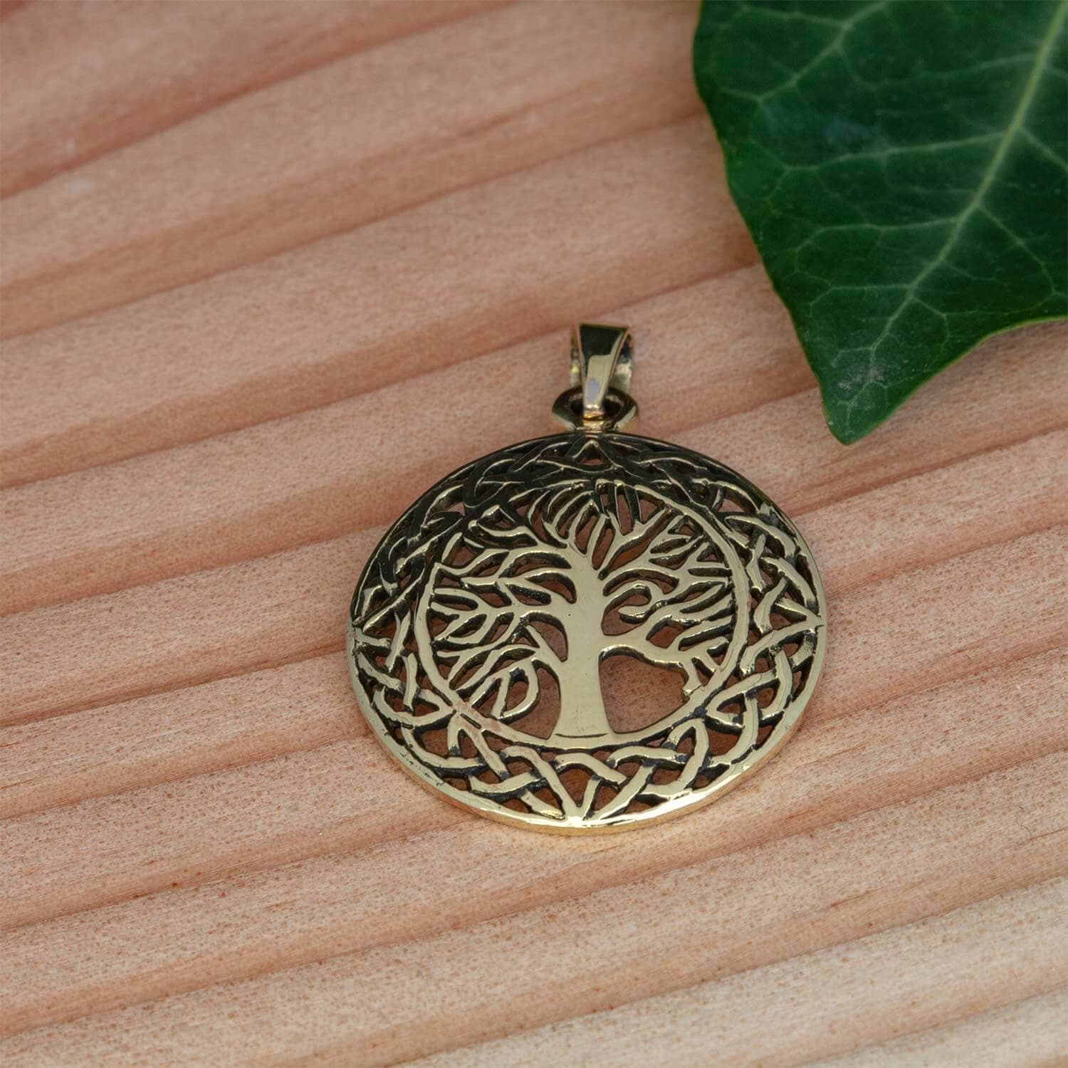 Et rundt Yggdrasil Livets Træ Vedhæng Qath-vedhæng i bronze med et indviklet, udskåret design af Livets træ i en keltisk knudecirkel, hvilende på en træoverflade. Et grønt efeublad er delvist synligt i øverste højre hjørne.