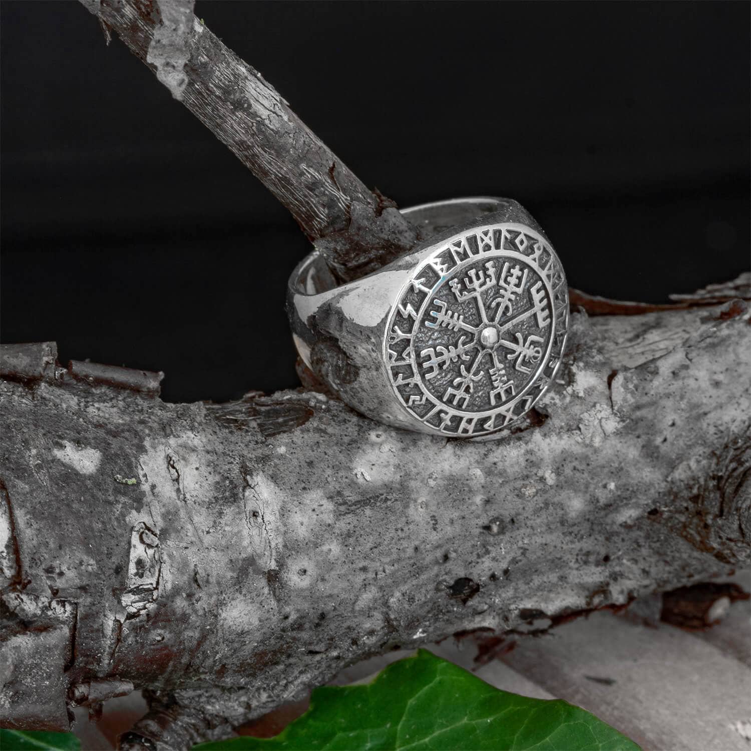 Sølvring Vejviser 925s Sølv, prydet med runesymboler, herunder en Vegvísir-rune, hviler på en ru trægren. Ringens detaljerede graveringer er lavet af 925'er sterlingsølv og skiller sig ud mod den mørke baggrund, og et grønt blad er synligt i nederste højre hjørne - et nik til nordisk mytologi.