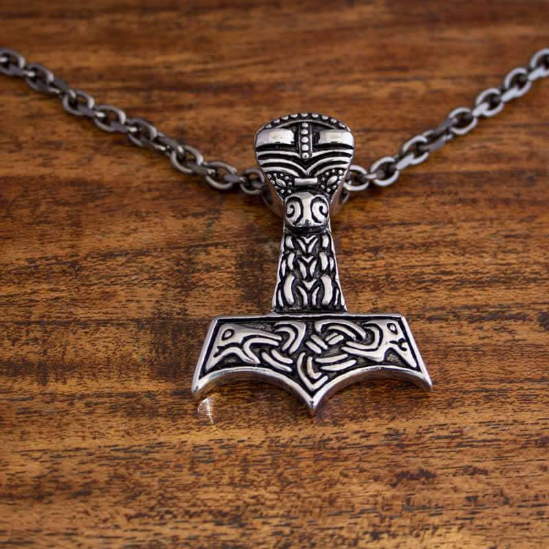 En sølvvedhæng med designet af Thors hammer, Mjolnir, med indviklede nordiske udskæringer og symboler på. Vedhænget er fastgjort til en Halskæde Ankerkæde Metallisk Stål Sort 5mm og hviler på en træoverflade.