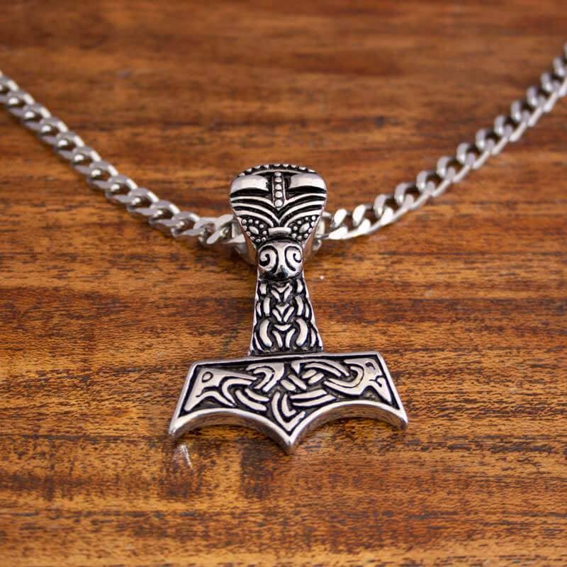 Et sølvvedhæng formet som Thors hammer, Mjölnir, hviler på Halskæde Panserkæde Stål 6mm, tilgængelig i forskellige længder. Hammeren er indviklet designet med detaljerede nordiske mønstre. Baggrunden har en træoverflade med en rig, varm tone.