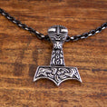 Bild in Galerie-Betrachter laden, Et detaljeret vedhæng i form af Thors hammer, Mjölnir, hænger fra en Halskæde Ankerkæde Sort Stål 5mm. Vedhænget har indviklede, nordisk-inspirerede udskæringer og mønstre i en slående kontrastfarve. Den hviler på en træoverflade.
